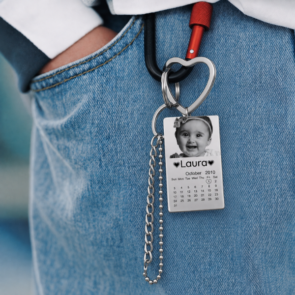 Gepersonaliseerde foto-kalendersleutelhanger aan een jeanslus, met een babyfoto, een gegraveerde naam en een kalenderdatum. Vaderdag cadeau, sentimenteel.